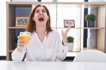 Brunette woman drinking glass of orange juice crazy and mad shouting and yelling with aggressive expression and arms raised. frustration concept.