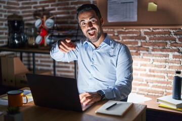 Hispanic man with beard working at the office at night pointing to you and the camera with fingers, smiling positive and cheerful