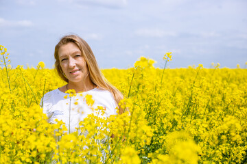 Naklejka na ściany i meble A blonde woman in a rapeseed field smiling. The arrival of spring and warmth