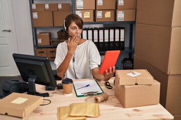 Brunette woman working at small business ecommerce with tablet covering mouth with hand, shocked and afraid for mistake. surprised expression