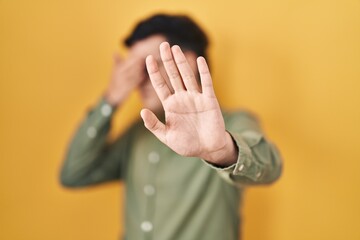 Non binary person standing over yellow background covering eyes with hands and doing stop gesture with sad and fear expression. embarrassed and negative concept.