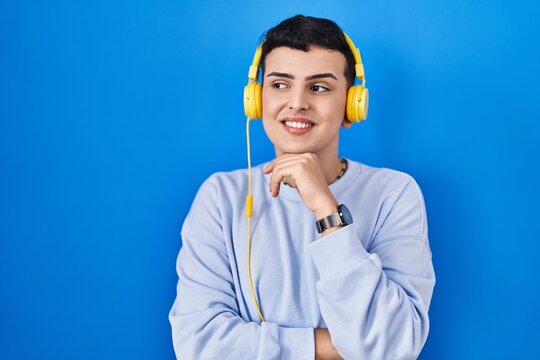 Non binary person listening to music using headphones with hand on chin thinking about question, pensive expression. smiling and thoughtful face. doubt concept.