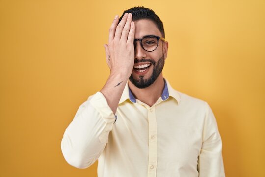 Hispanic young man wearing business clothes and glasses covering one eye with hand, confident smile on face and surprise emotion.