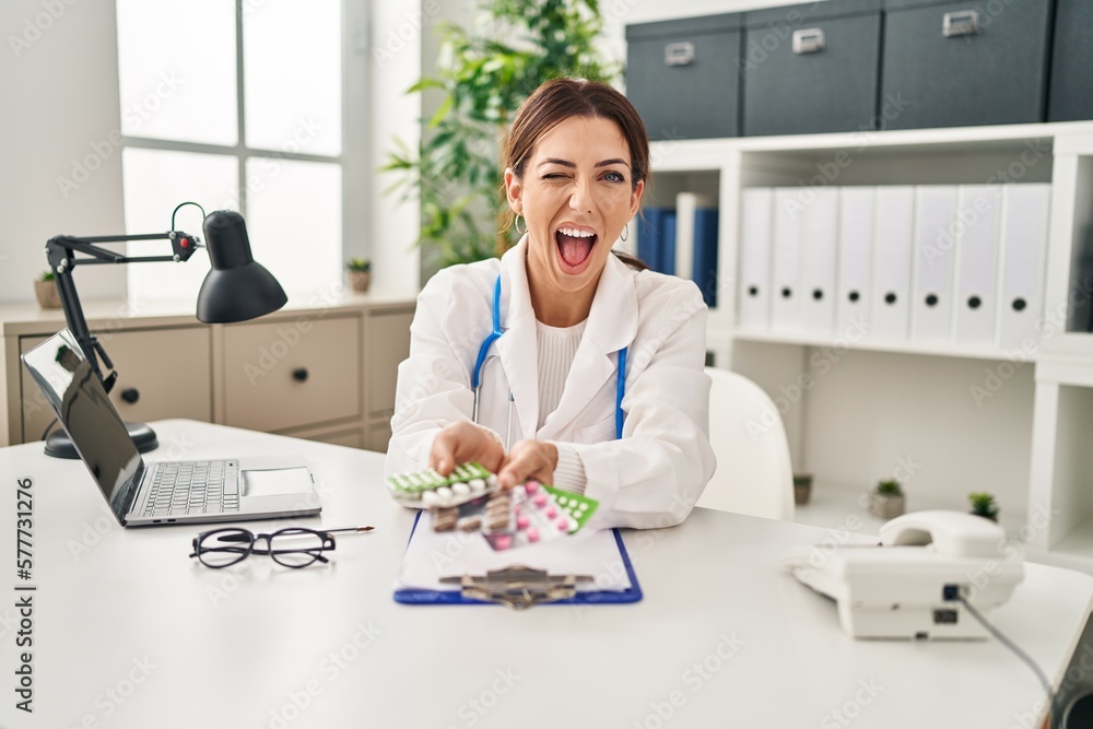 Sticker Young brunette doctor woman holding pills winking looking at the camera with sexy expression, cheerful and happy face.