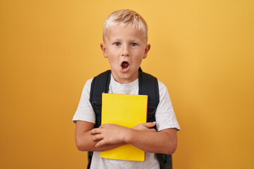 Little caucasian boy wearing student backpack and holding book scared and amazed with open mouth for surprise, disbelief face