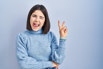 Young hispanic woman standing over blue background smiling with happy face winking at the camera doing victory sign with fingers. number two.