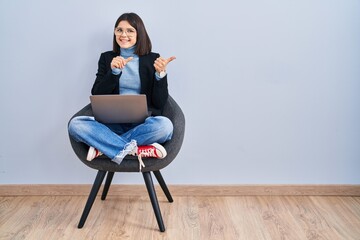 Young hispanic woman sitting on chair using computer laptop pointing to the back behind with hand and thumbs up, smiling confident