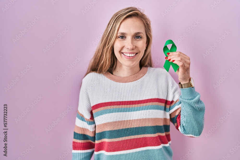 Sticker Young blonde woman holding support green ribbon looking positive and happy standing and smiling with a confident smile showing teeth