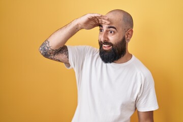 Fototapeta na wymiar Young hispanic man with beard and tattoos standing over yellow background very happy and smiling looking far away with hand over head. searching concept.
