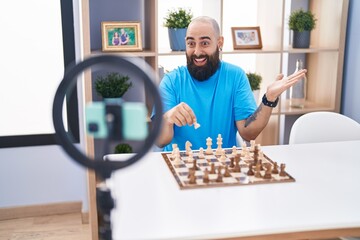 Young hispanic man with beard and tattoos recording chess tutorial with smartphone at home screaming proud, celebrating victory and success very excited with raised arm