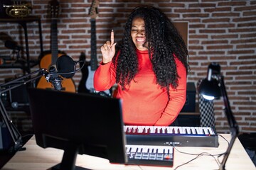 Plus size hispanic woman playing piano at music studio showing and pointing up with finger number one while smiling confident and happy.