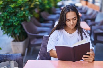 Young beautiful hispanic woman reading book sitting on table at coffee shop terrace