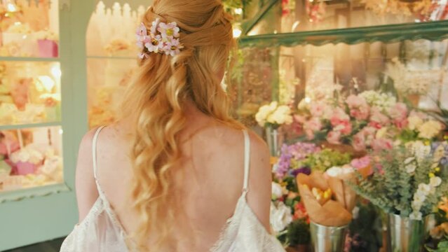 Portrait blonde woman in white dress style florist seller looking at shop window. girl fashion model stylish hairstyle curly blond hair with pink flowers back view. Spring street Lady chooses flowers