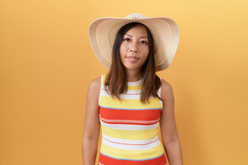 Middle age chinese woman wearing summer hat over yellow background looking sleepy and tired, exhausted for fatigue and hangover, lazy eyes in the morning.