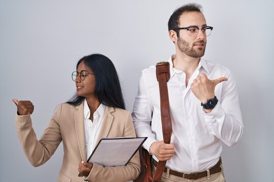 Interracial business couple wearing glasses smiling with happy face looking and pointing to the side with thumb up.