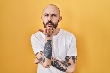 Young hispanic man with tattoos standing over yellow background looking at the camera blowing a kiss with hand on air being lovely and sexy. love expression.