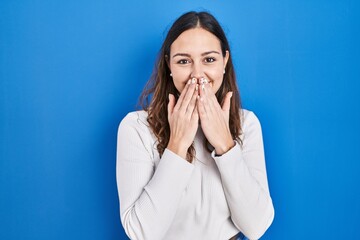 Young hispanic woman standing over blue background laughing and embarrassed giggle covering mouth...