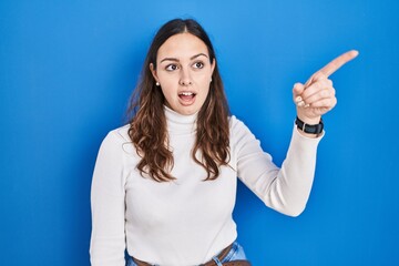 Young hispanic woman standing over blue background pointing with finger surprised ahead, open mouth amazed expression, something on the front