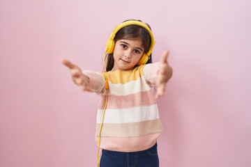 Little hispanic girl listening to music using headphones looking at the camera smiling with open arms for hug. cheerful expression embracing happiness.