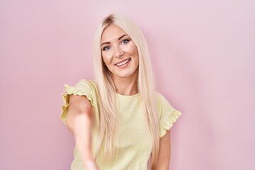 Caucasian woman standing over pink background smiling friendly offering handshake as greeting and welcoming. successful business.