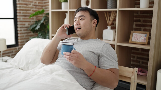 Young chinese man talking on smartphone drinking coffee at bedroom