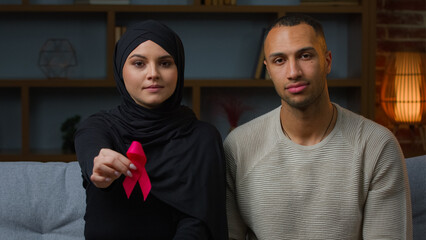 Multiracial family muslim woman and african american man sit on sofa indoors hold red ribbon...