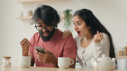 Multiracial couple indian man and woman sit in kitchen using mobile phone application enjoying web...