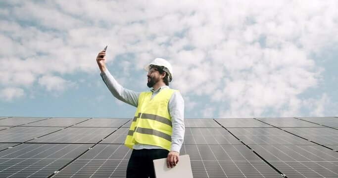 Male engineer speaking on smartphone on solar farm. Low angle man in uniform walking near photovoltaic panels and answering phone call against cloudy blue sky on sunny day on solar power station.