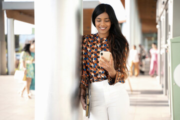 Beautiful smiling Indian woman holding smartphone using mobile app shopping online on the street. Modern asian female looking at mobile phone screen, reading text message, chatting. Mobile banking