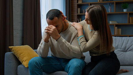 Offended African american man sit on couch upset with bad news health problem Caucasian woman consoling apologize ask forgiveness after quarrel wife touching husband calming support family conflict 