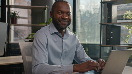 Professional middle-aged mature 40s ethnic African American man senior businessman office worker sit indoors search internet data on laptop pc device looking at camera friendly smiling positive smile