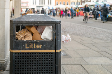 Shallow focus of a typical city litter bin showing it full of rubbish. A nearby English market is seen.