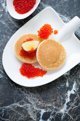 Shovel-shaped plate with red caviar and mini pancakes, high angle view on a black marble background, vertical shot