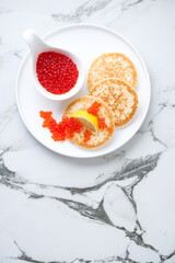 Mini pancakes with red caviar on a white marble background, vertical shot, copy space, flat lay