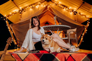 Happy young woman with her Welsh Corgi Pembroke dog relaxing in glamping on summer evening near cozy bonfire. Luxury camping tent for outdoor recreation and recreation. Lifestyle concept - 577745207