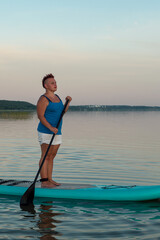 A woman in mohawk shorts stands on a SUP board with a paddle at sunset in a lake against a pink-blue sky.