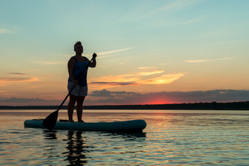 A woman in shorts and a T-shirt with a Mohawk stands on a SUP board in the evening at sunset in the lake.