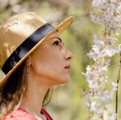 A young beautiful woman close-up in a straw hat admires the white bright cherry blossoms in the spring garden