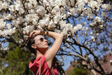 A beautiful young woman in a red dress and sunglasses admires a white flowering magnolia in a spring garden