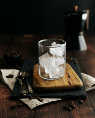 Taza de cristal transparente con hielo sobre una tabla de madera, jarra con café y fondo negro