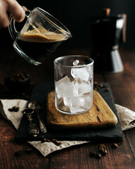 Jarra de café siendo servido en una taza de cristal transparente con hielo sobre una tabla de madera y fondo negro