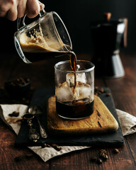 Jarra de café siendo servido en una taza de cristal transparente con hielo sobre una tabla de madera y fondo negro