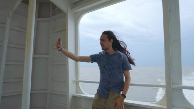 Young latin man on the deck of a ferry taking a selfie while the wind blows his hair. 4k video.