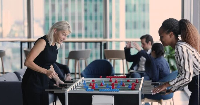 Office team playing table foosball relaxing at lunch break in modern office. Two multiethnic happy female employees have fun in workplace, relieving stress at workday, enjoy free time with colleague