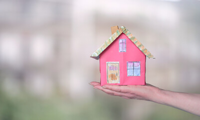 Construction and architecture concept, cute model home on a female hand on blurred buildings background