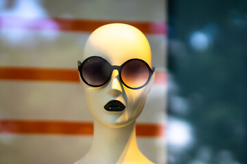 A bald doll's head with black painted lips and dark sunglasses behind a shop window