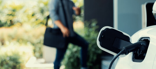 Focus electric car charging at home charging station with blurred progressive man walking in the...