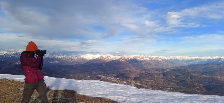 Young caucasian woman using DSLR camera to take a photo from the top of Ceuze mountain during winter. View on the valley and the city of Gap. She wear winter sport clothes.