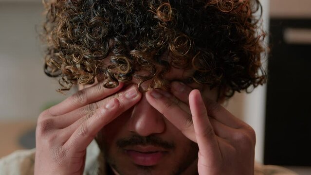 Close up Indian ill tired sleepy man with curly hair rubbing dry eyes Arabian exhausted sick guy businessman indoors suffer rub eye strain discomfort headache bad eyesight astigmatism ophthalmology