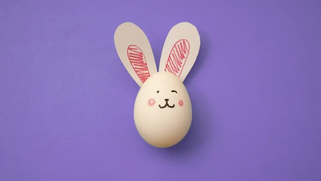 Top view 4K animated video. Frame-by-frame, stop motion animation. White chicken egg with cute bunny face and rabbit ears on purple background. Postcard for religious holiday Happy Easter. Magic. Food